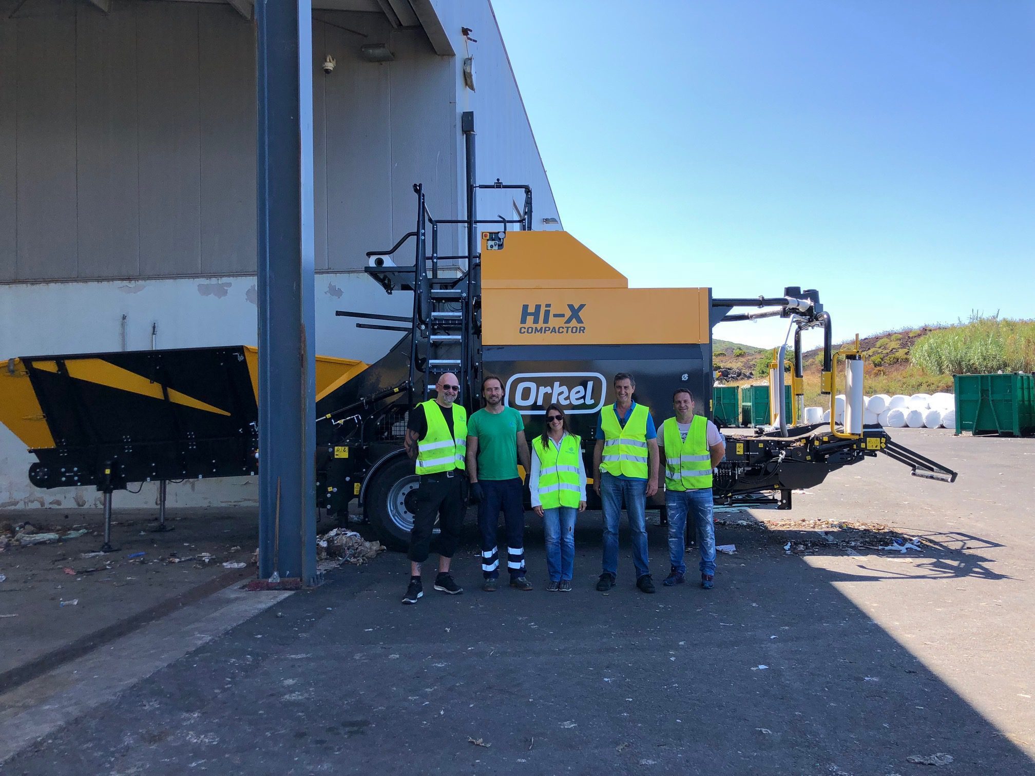 Fünf Personen stehen im Schatten einer Hi-X-Ballenpresse mit blauem Himmel und weißen Rundballen im Hintergrund.