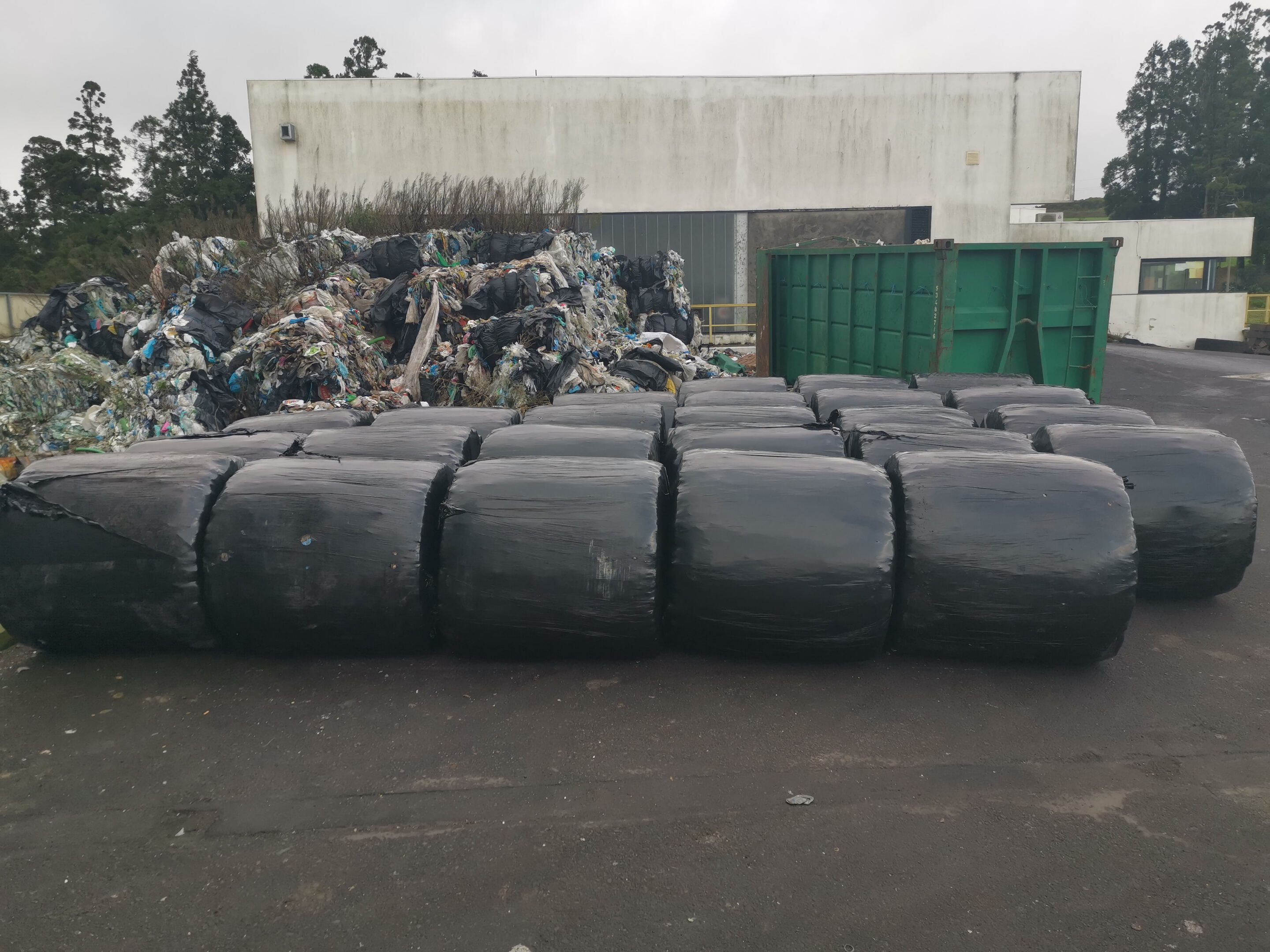 Schwarze Rundballen liegen auf ihrer runden Seite mit einem Haufen farbigen Deponiematerials im Hintergrund.