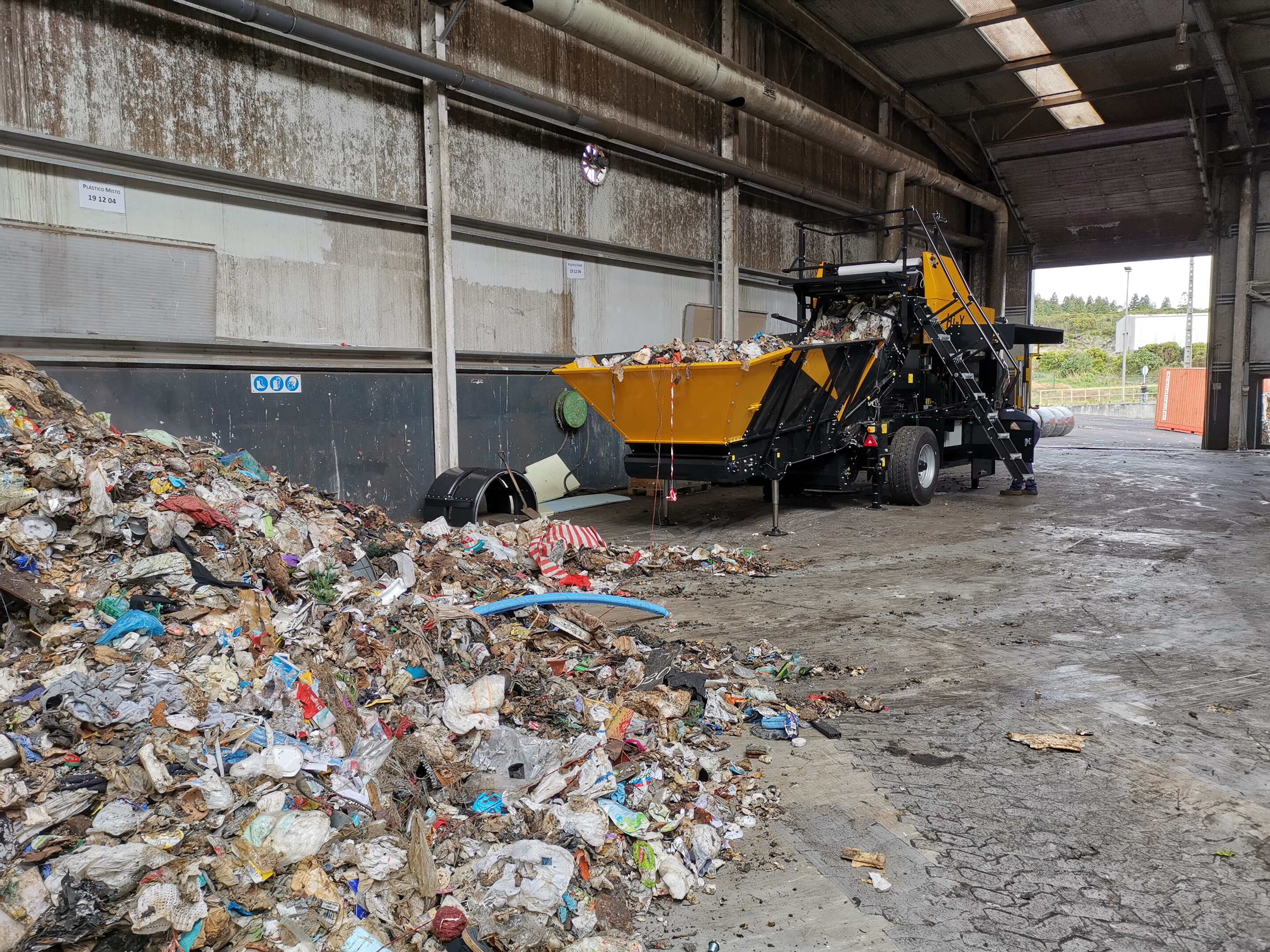 Eine Hi-X-Ballenpresse in einer Betonanlage, mit losem Deponieabfall in ihrem Zuführtisch und Haufen von losem Abfall im Vordergrund.