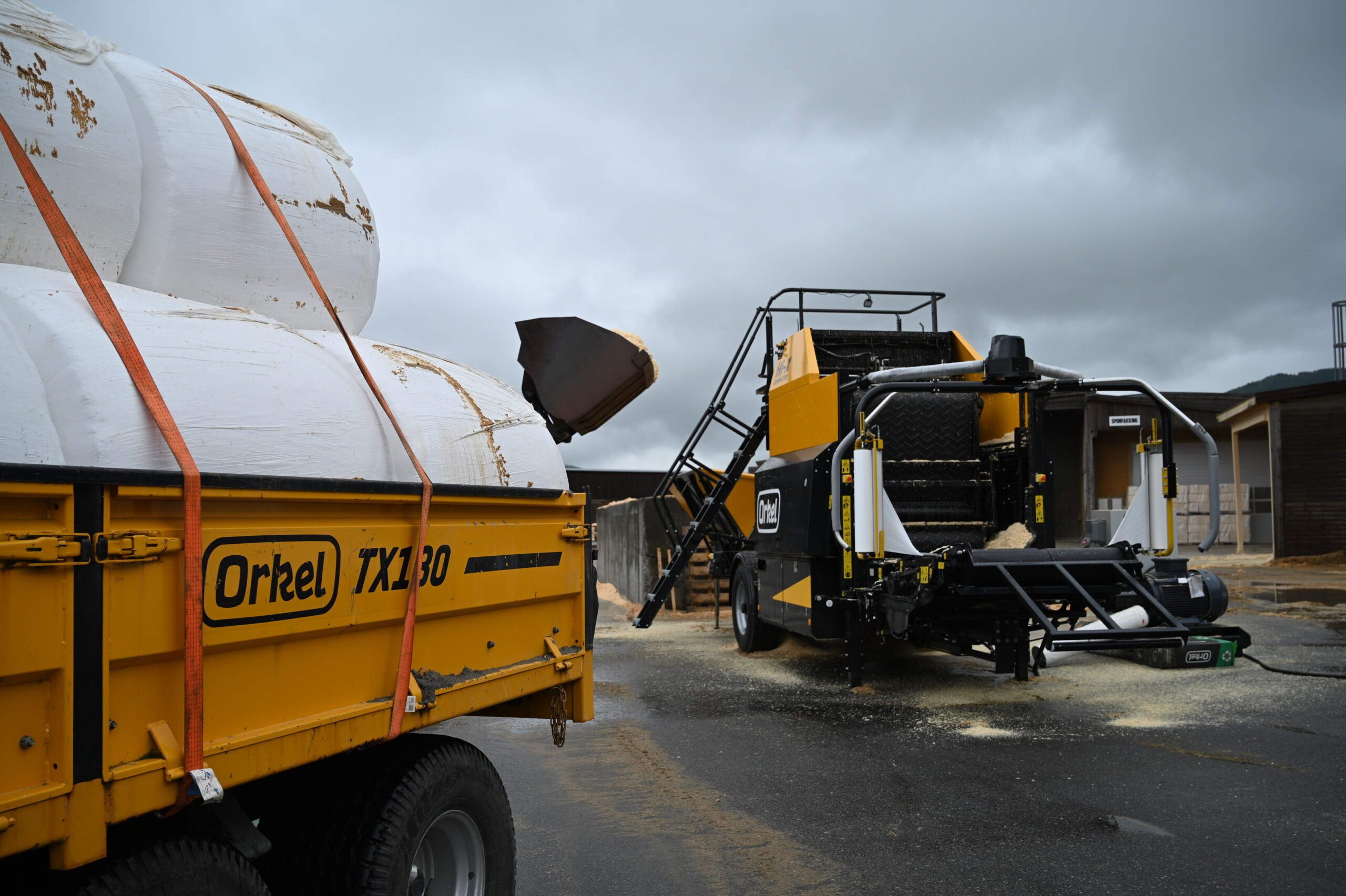 A Hi-X compactor in the background, with a yellow trailer filled with round bales in the foreground.