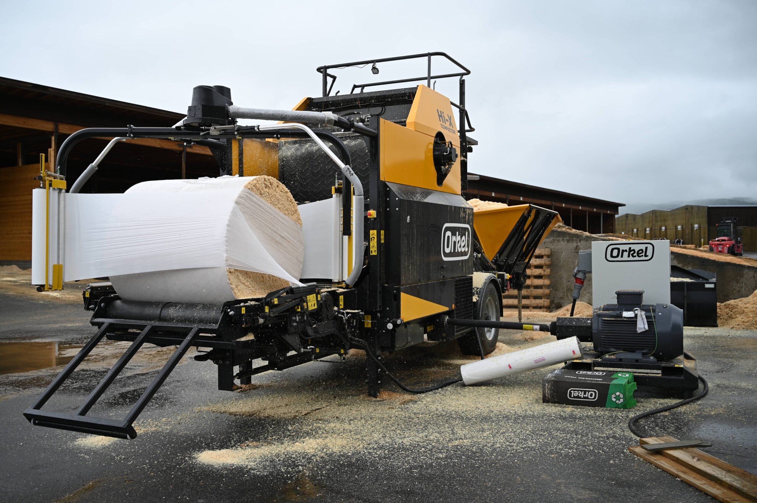 A Hi-X compactor with a half-wrapped wood shavings bale on its wrapping table.