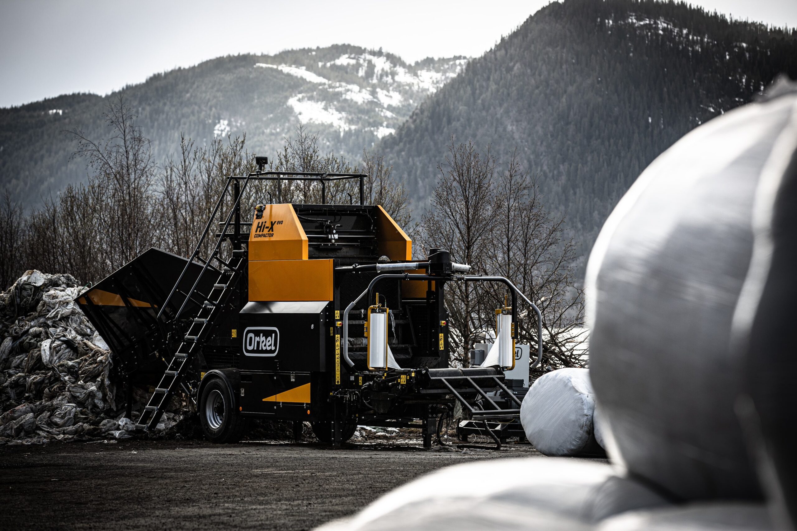 The Hi-X evo compactor with white skies and hills in the background and blurry round bales in the foreground.