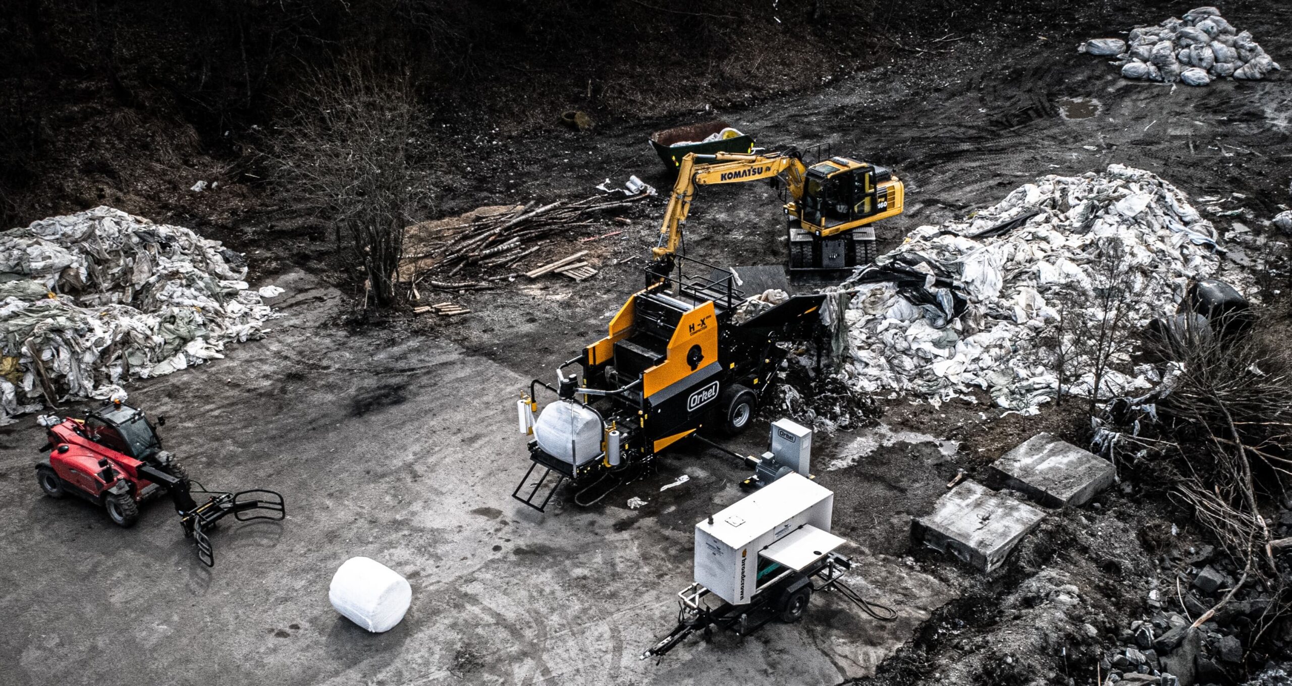 Overhead shot of a Hi-X evo baling a large pile of plastic waste, with a yellow excavator above and a bale grabber to the left.
