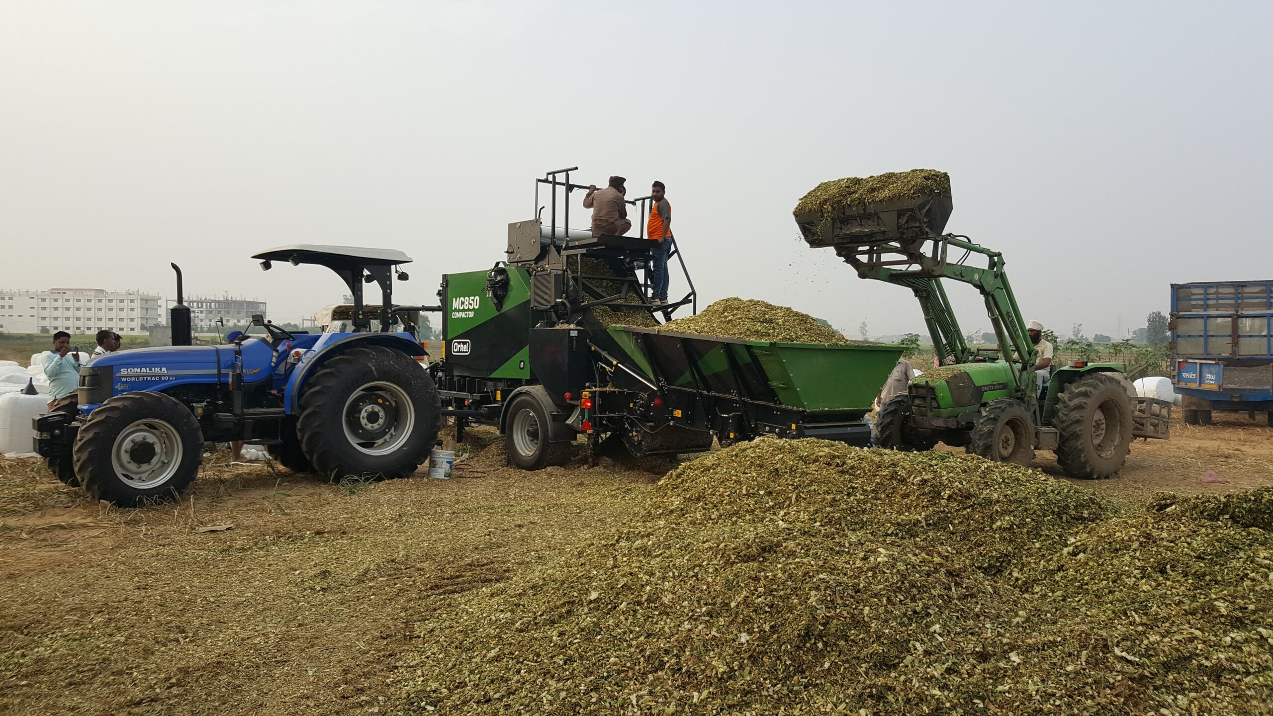 An MP850 compactor baling, with a tractor dumping bulk material into the machine's feed hopper and large quantities of material in the foreground.