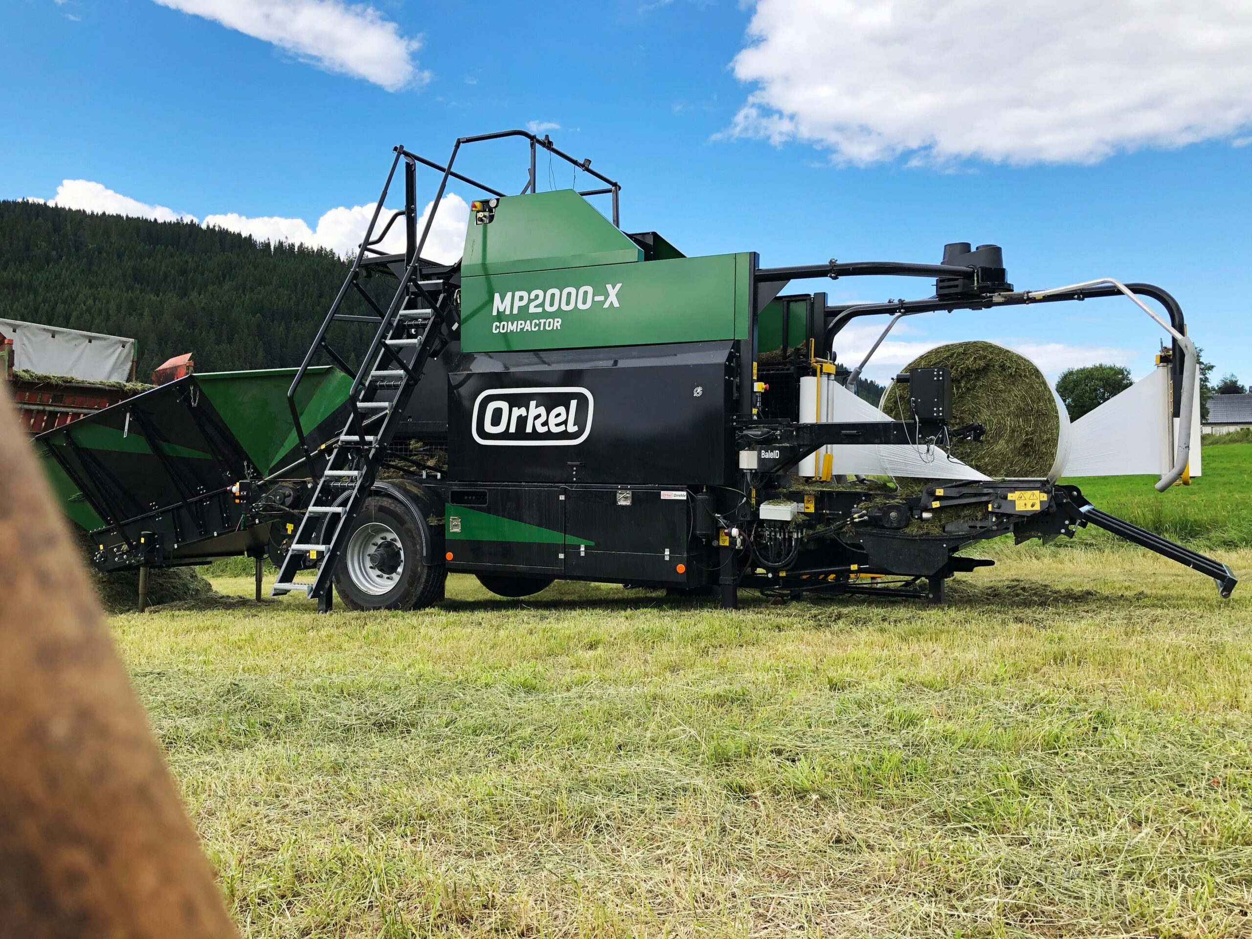 An MP2000-X in a green field with a blue sky, in the middle of wrapping a grass bale.