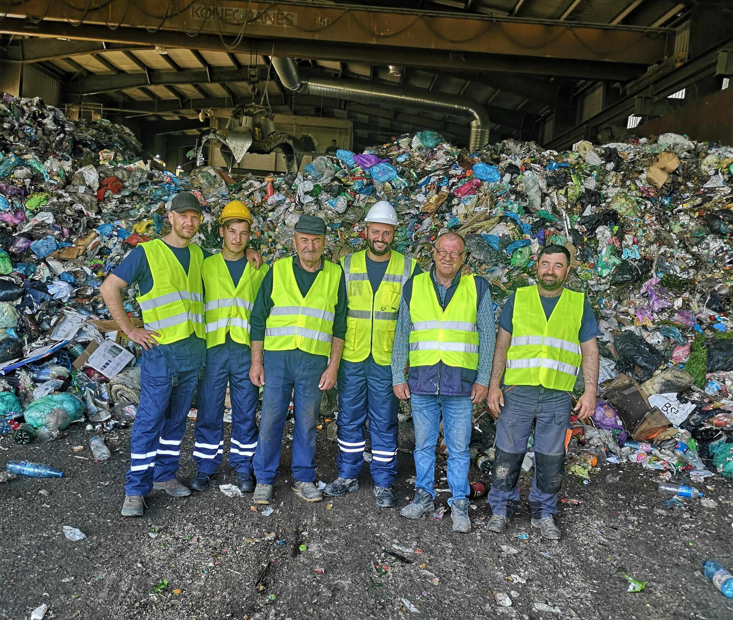Seis pessoas com coletes de segurança amarelos posando em frente a uma pilha alta de resíduos domésticos, trabalhando na gestão de resíduos