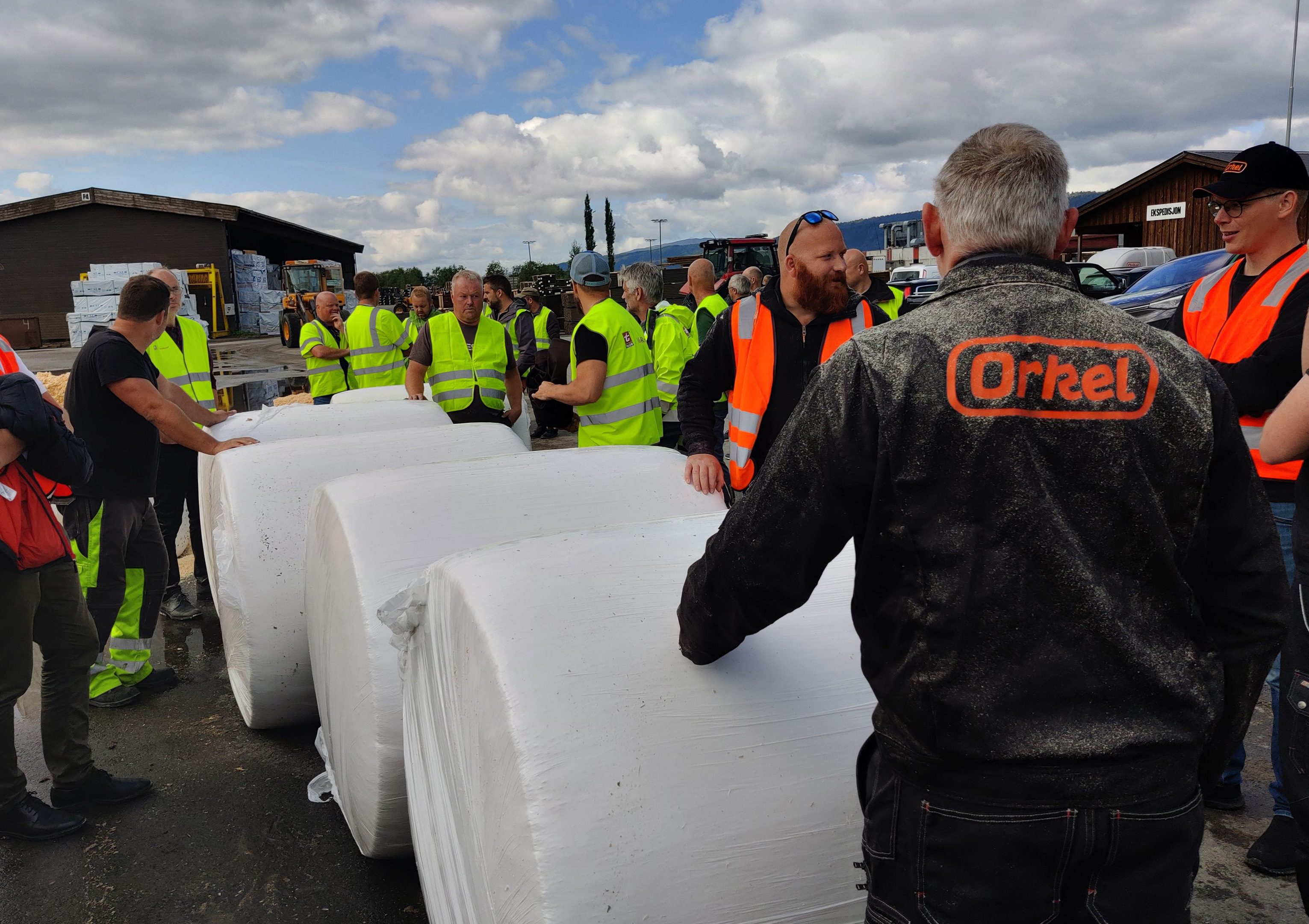 Zahlreiche Personen in gelben oder orangefarbenen Sicherheitswesten stehen um weiße Rundballen herum.