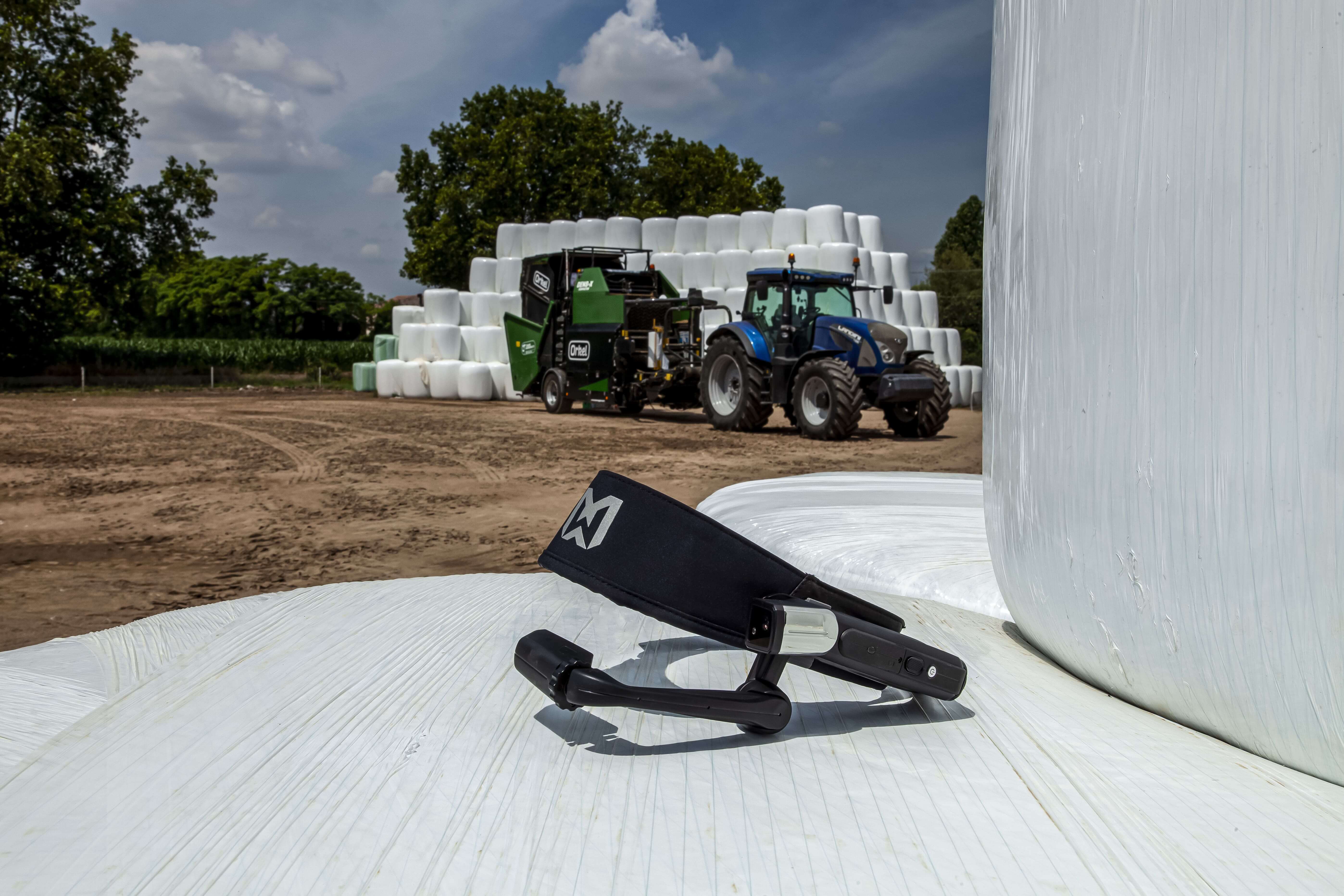 Closeup on a realwear device with an Orkel compactor and white round bales in the background