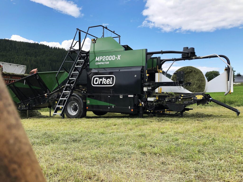 A green Orkel compactor with a round bale on the wrapping table, standing on a field in sunny weather, baling short chopped grass