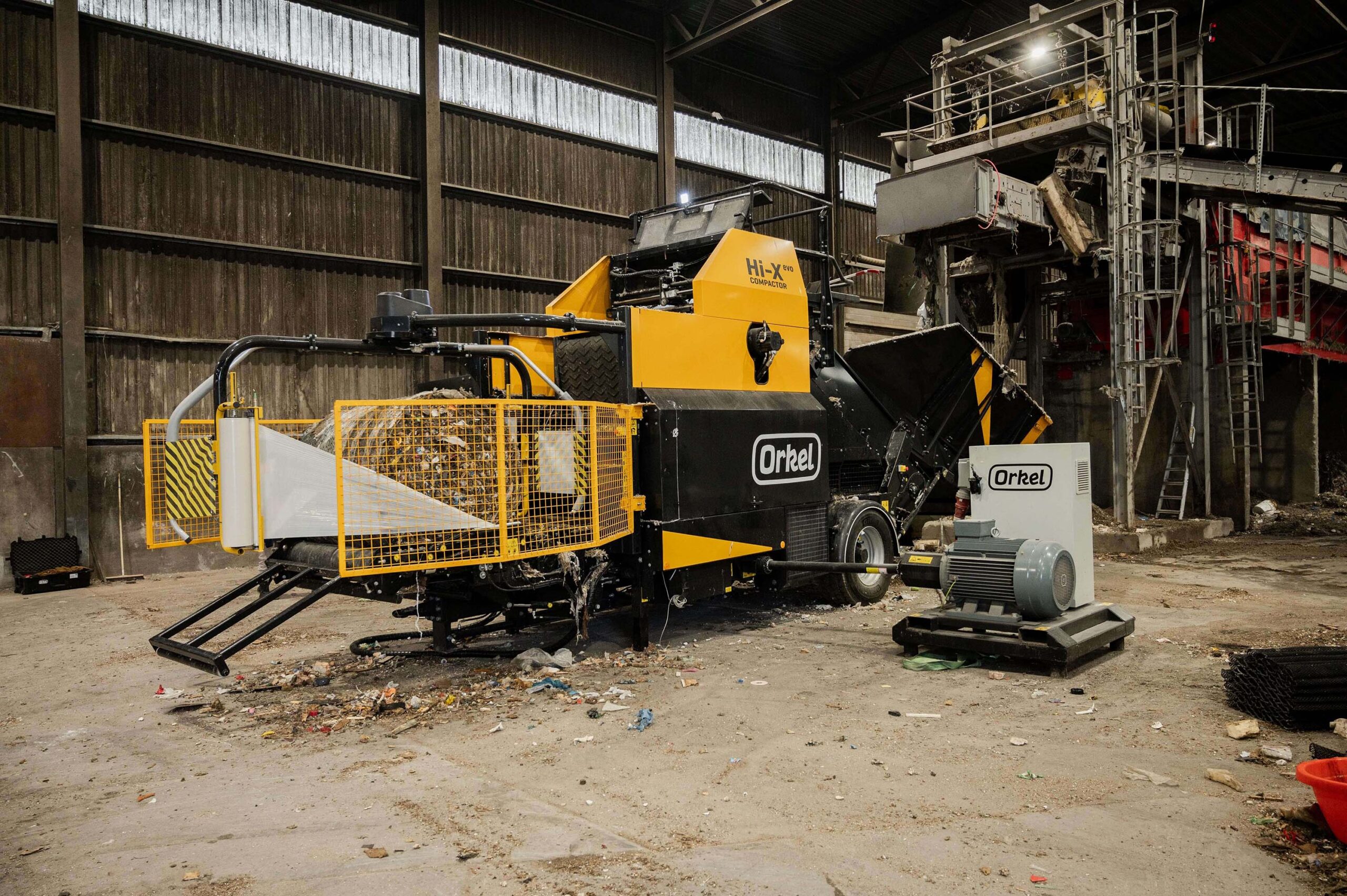 A yellow Orkel compactor standing under a waste shredder in a waste facility