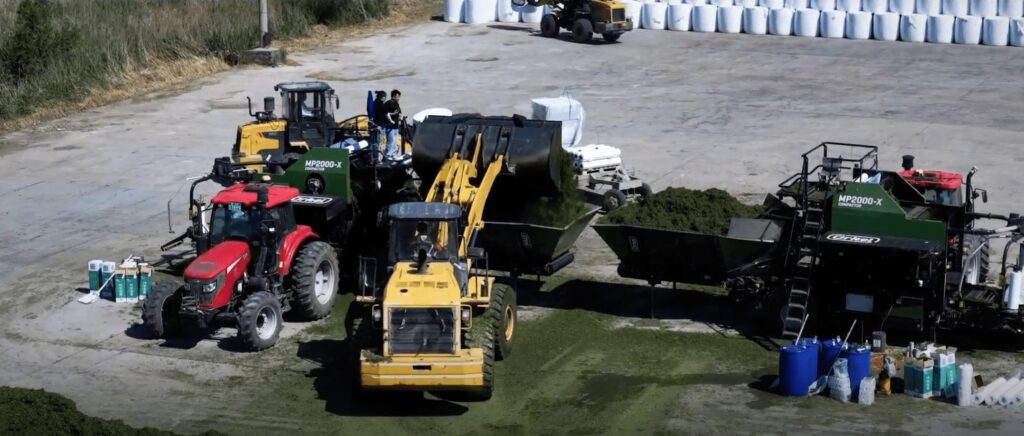 Dois compactadores agrícolas a enfardar numa grande área de betão com fardos redondos ao fundo. Um carregador frontal despeja material nas tremonhas de alimentação 