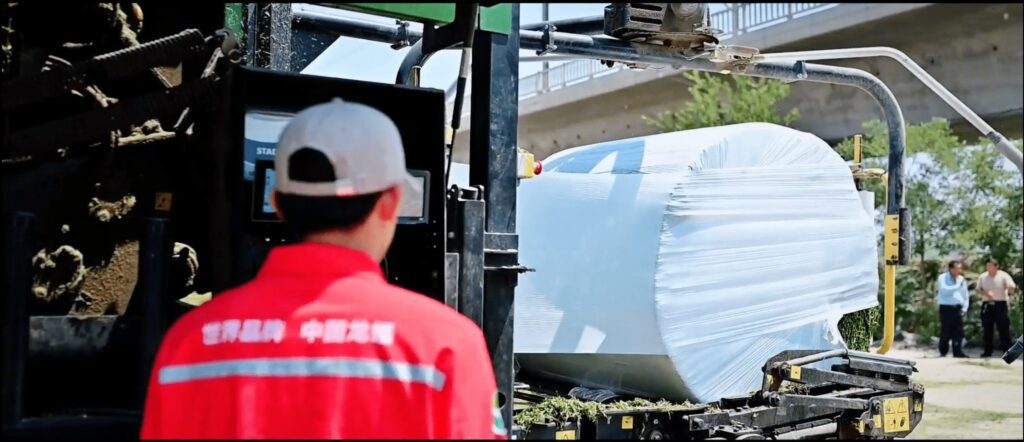 As costas de um homem usando um casaco laranja e um chapéu branco de sol em primeiro plano, olhando para um compactador agrícola a envolver um fardo redondo