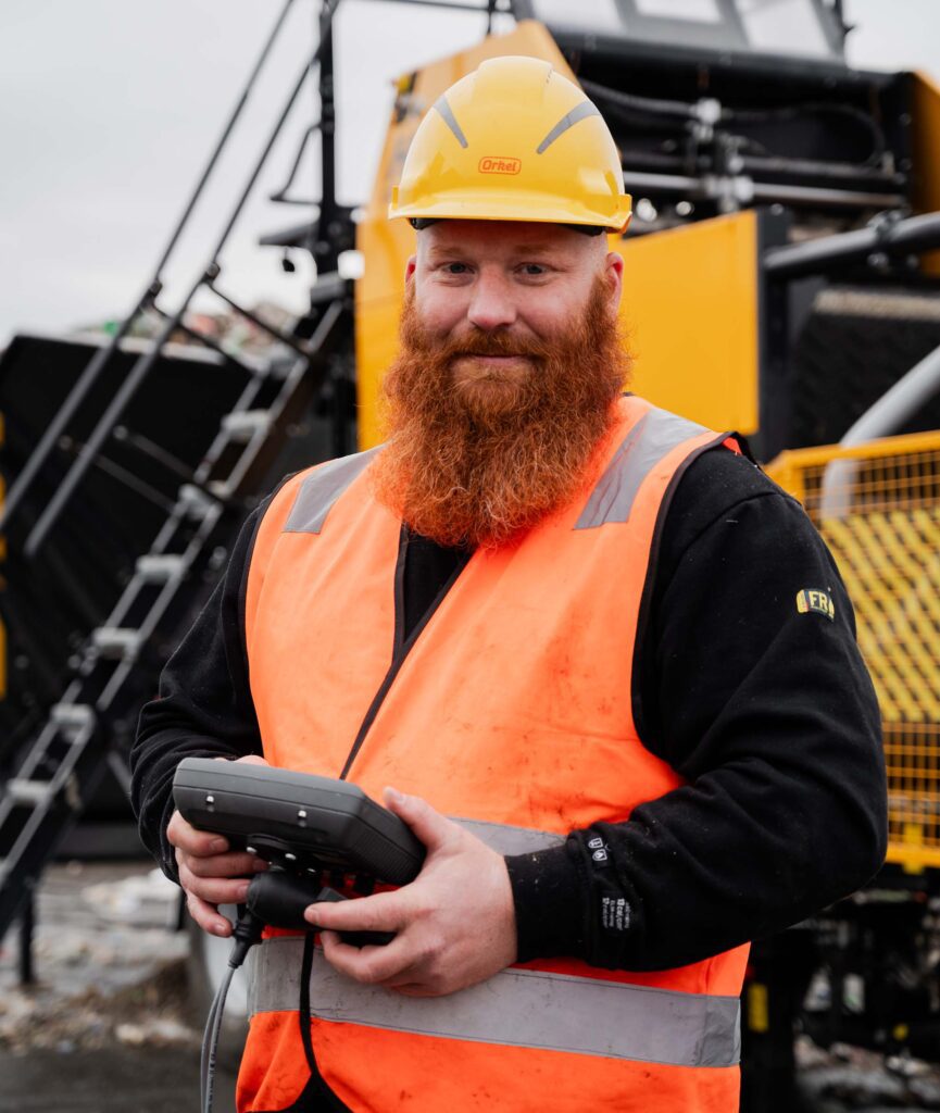 Ein Mann in Sicherheitsweste und Helm steht selbstbewusst in einer Abfallanlage, mit einer großen Industriemaschine im Hintergrund. Die Umgebung umfasst Stapel von recycelbaren Materialien und schweres Gerät, was eine industrielle Umgebung mit Fokus auf Abfallmanagement und -verarbeitung unterstreicht.