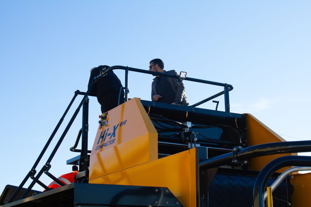 Zwei Männer stehen auf der Plattform einer industriellen Ballenpresse und blicken von der Kamera weg.