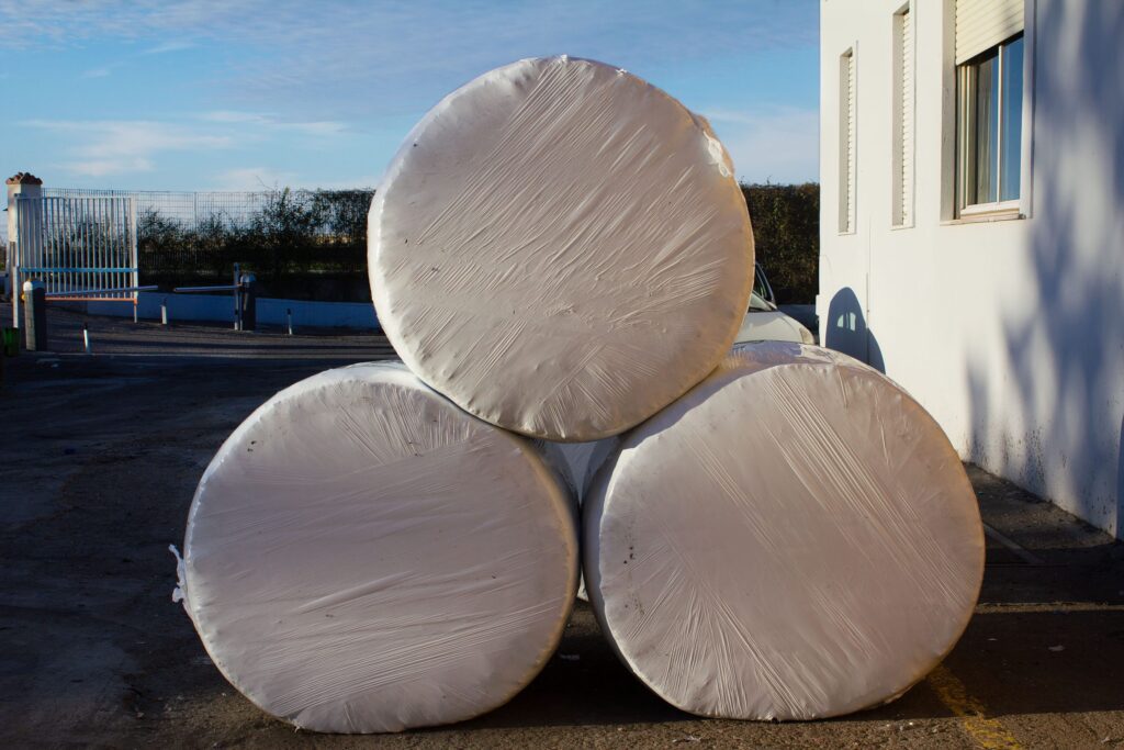 Drei in weißes Plastik gewickelte Rundballen, ordentlich neben einem weißen Gebäude gestapelt.