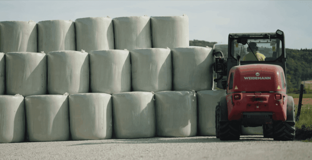 Ein ordentlicher Stapel Rundballen, drei Stockwerke hoch, und ein Gabelstapler, der von hinten einen Ballen in den Stapel legt.