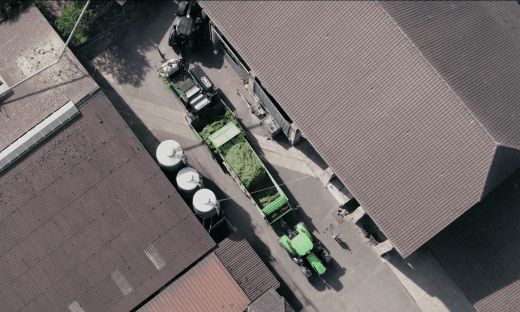 Von oben gesehen, eine landwirtschaftliche Presse, die Rundballen herstellt, und ein Traktor mit einem Anhänger, der Gras in die Presse entlädt. Auf der linken und rechten Seite sehen wir Hausdächer.