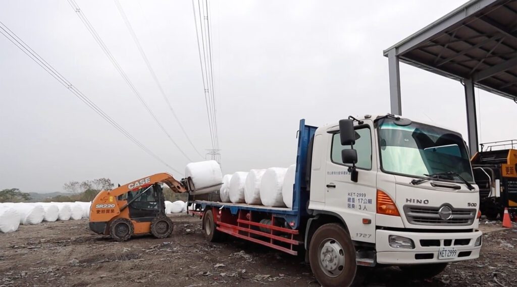 Un chariot élévateur chargeant des balles rondes sur un camion pour le transport, à l'extérieur d'un grand hangar