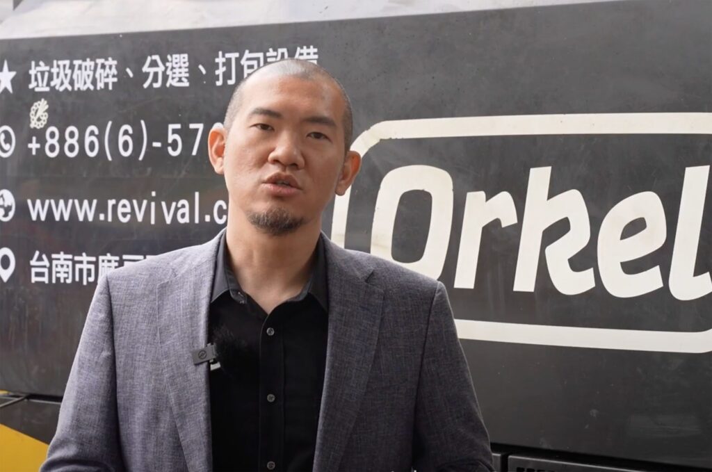 Un homme interviewé, portant un costume et un polo noir. Une machine industrielle en arrière-plan