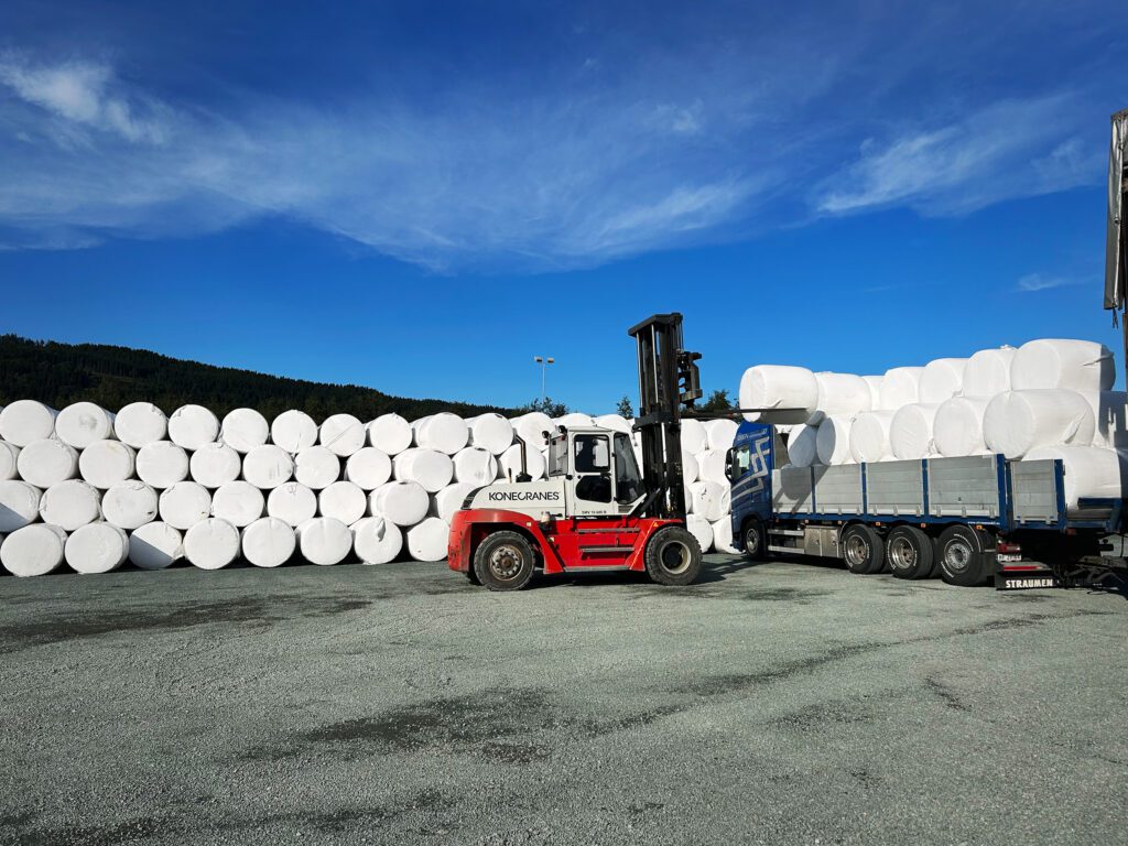 Forklift loading a round bale onto a trailer full of round bales. Stacks of round bales in the background and blue skies